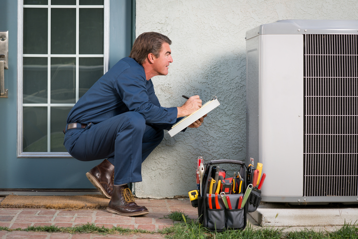 Air conditioner repairman in uniform with a clipboard and tool bucket inspecting the AC unit. | Advanced Heating and Air Conditioning